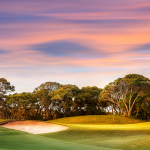 A serene golf course at dusk with a vibrant, painted sky in shades of pink and purple. The well-maintained green is bordered by a sand bunker on the left and a cluster of trees providing a natural backdrop. A single flag marks the hole, offering an idyllic setting for golf enthusiasts.