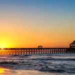 A serene sunset over the ocean with the sun hovering just above the horizon, casting a warm golden glow across the sky and water. A silhouette of a long pier at Folly Beach with intricate lattice patterns extends into the sea, featuring a small gazebo-like structure at its far end. Gentle waves lap at the sandy shore in the foreground, reflecting the sunset’s light. The scene conveys a sense of calm and natural beauty