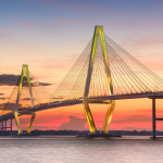 A panoramic view of the Ravenel Bridge at sunset, with vibrant hues of pink and orange in the sky. The bridge features distinctive cable-stayed construction with two tall, illuminated towers and numerous converging support cables. The calm water below reflects the warm glow of the lights on the bridge’s underside, and Mount Pleasant is visible in the background under a serene twilight sky.