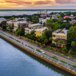 Aerial view of a Charleston at dusk with a clear view of a long waterfront street lined with historic buildings. The calm water reflects the soft colors of the sky, and lush greenery is interspersed among the structures. The image captures the tranquil beauty of the city’s interface with nature.