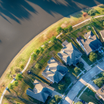 Aerial view of a residential neighborhood adjacent to a body of water, casting long shadows in the low sunlight. The neighborhood consists of several houses with dark roofs, arranged neatly around a curved street that follows the shoreline. The houses have well-maintained lawns and driveways connected to the street. The water body reflects the sunlight, creating a bright glint on its surface. The contrast between the built environment and natural water feature creates an interesting visual dynamic.