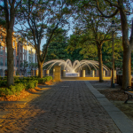 A serene park scene with a cobblestone pathway leading towards a central fountain. The fountain has multiple streams of water arching into the air, creating a symmetrical pattern. Surrounding the fountain are several wooden benches and lush green trees, casting dappled shadows on the ground. In the background, there are multi-story buildings with numerous windows reflecting the early morning or late afternoon sunlight. The sky is clear with a soft gradient from light blue to warm orange hues near the horizon, suggesting either sunrise or sunset. This scene is reminiscent of North Charleston Riverfront Park.