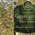 Welcome to Charleston West Ashley” sign, featuring an emblem and the subtitle “Birthplace of Charles Towne” against a backdrop of lush green trees. The sign is mounted on a metal post and is prominently displayed in a natural setting with dappled sunlight filtering through the foliage, indicating a historic or significant location within the community